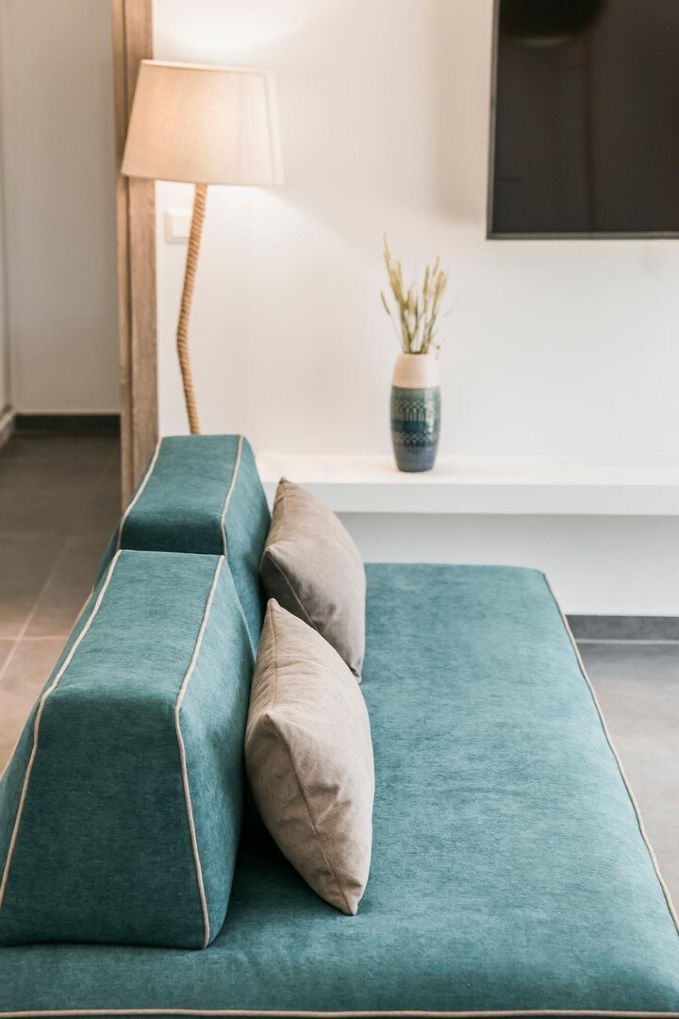 Ein Türkisblaues Sofa mit Holzrahmen und zwei Kissen, eine Stehlampe sowie eine Vase mit getrockneten Pflanzen auf einem weißen Regal.