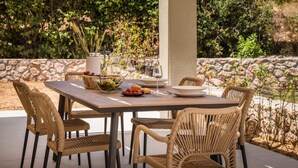 An outdoor dining area with a table set for four, surrounded by wicker chairs, with a stone wall and greenery in the background. - Villa Amarylis - Ionian Treasure Villas (Kefalonia)