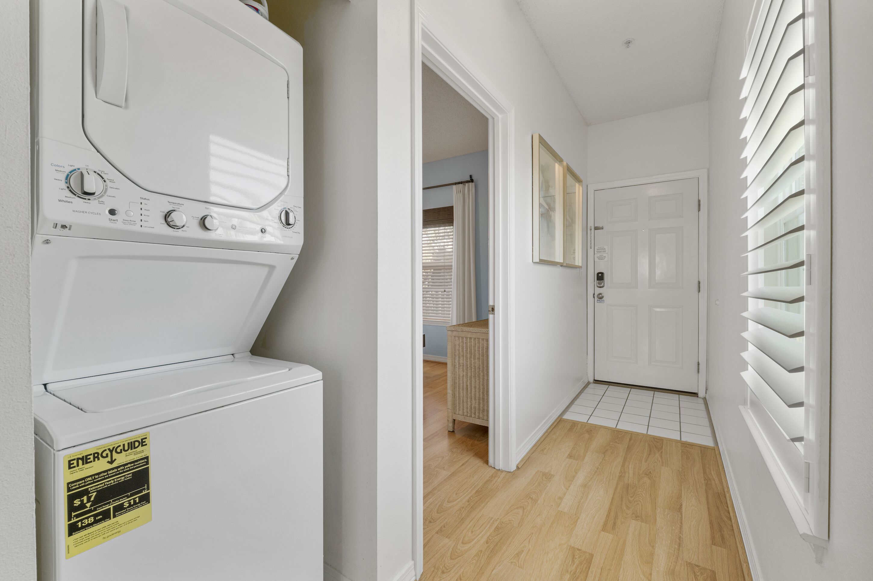 A laundry room with a front-loading washer and dryer, a door leading to a hallway, and a window with blinds.