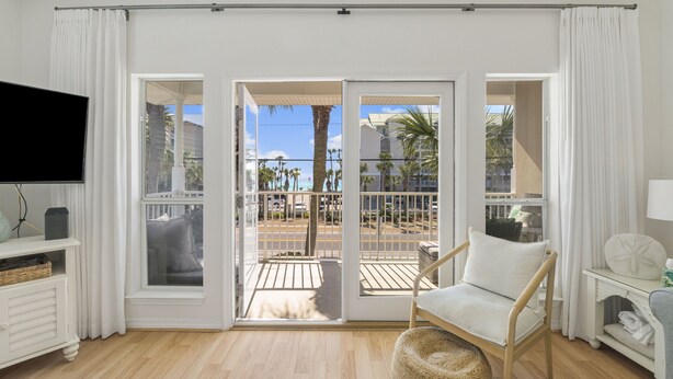 A room with a TV, a wicker chair, a small table, and a view of a balcony with a railing and palm trees. - Caribbean Dunes by Panhandle Getaways (Destin)