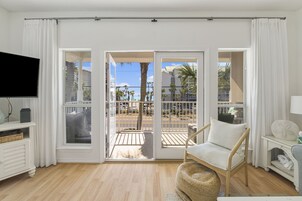 A room with a TV, a wicker chair, a small table, and a view of a balcony with a railing and palm trees.