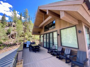 A modern outdoor deck with a wooden table and chairs, surrounded by large windows and a view of a mountainous landscape.