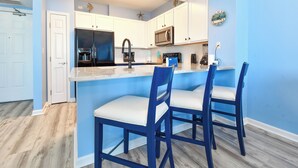 A modern kitchen with white cabinets, a black refrigerator, a microwave, and a coffee maker. There are two blue bar stools with white cushions. - Cozy condo w/ Vibrant Amenities & Zero-Entry Lagoon Pool (Destin)