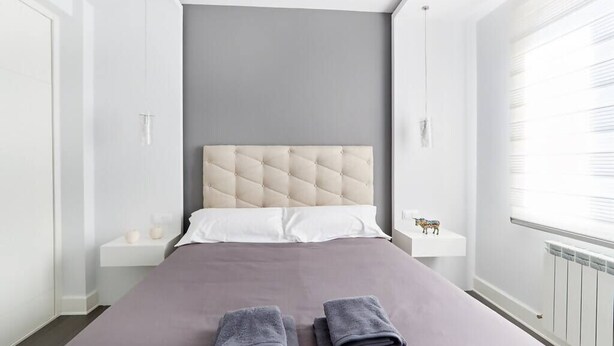 Un dormitorio moderno con cabecera capitonada, una paleta de colores neutros y una cama bien hecha con toallas dobladas.