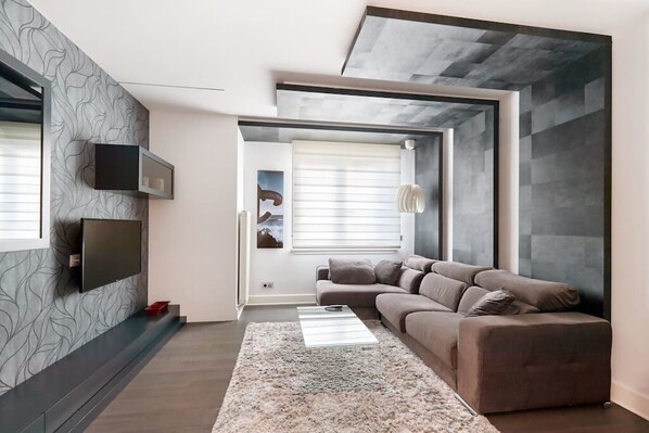 Una sala de estar moderna con una pared gris estampada, un sofá gris, una mesa de centro de vidrio y un ventanal con persianas.
