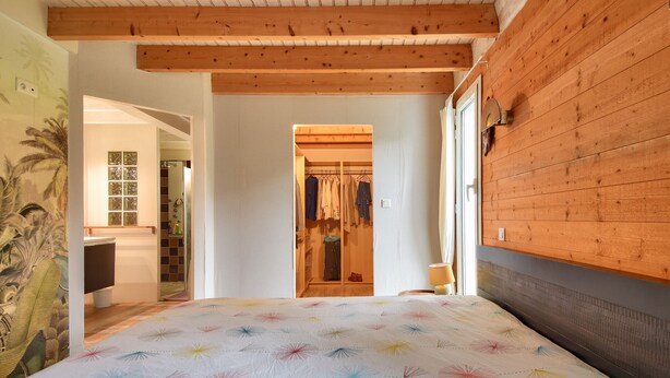 Ein Schlafzimmer mit Bett, Holzbalkendecke, einem Kleiderschrank mit Kleidung und Blick in einen weiteren Raum.