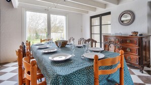 Ein Esszimmer mit einem langen Tisch, gedeckt für sechs Personen, Holzstühlen, einer gemusterten Tischdecke, einem Schrank mit Schubladen und einem großen Fenster, das für natürliches Licht sorgt.
