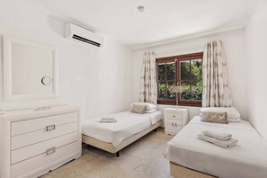 Een nette, witte kamer met twee eenpersoonsbedden, een dressoir, een raam met gordijnen en een airconditioning.