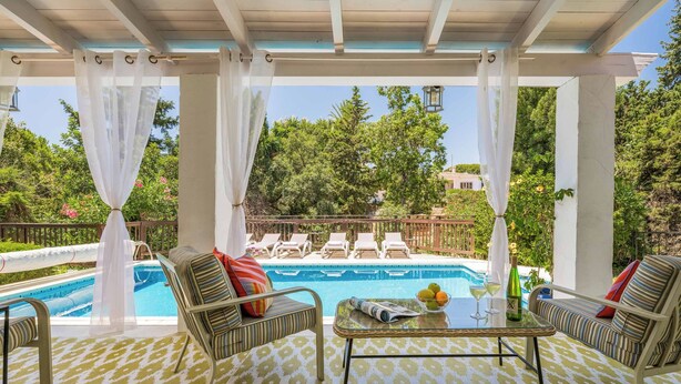 A covered outdoor seating area with a pool, lounge chairs, and a table with drinks and a newspaper.