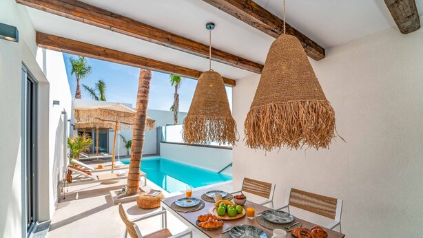 A dining area with a pool view, featuring a wooden table, wicker chairs, and a hanging light fixture.