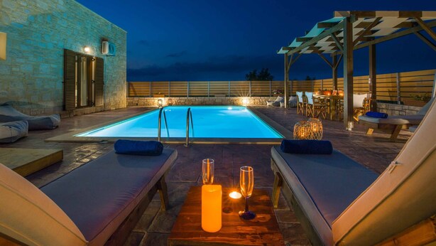 Un área junto a la piscina con sillones reclinables, una mesa de madera con velas, y un edificio de piedra al fondo.