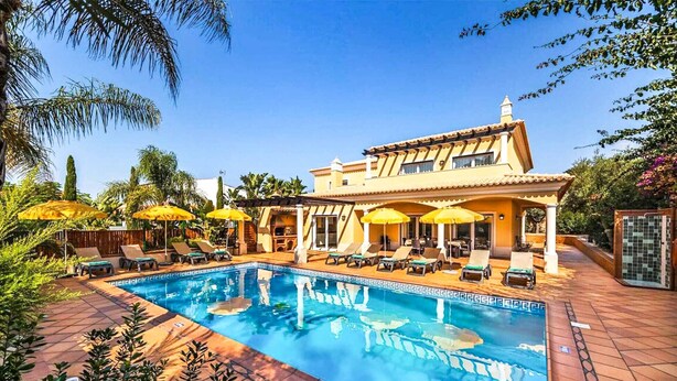 A pool area with lounge chairs and umbrellas, a pool with a tiled border, and a building with a tiled roof and windows. - Villa Cabrita de Parra (Vale Rebelho)