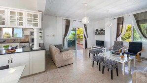A modern living room with a sofa, dining table, and chairs. There is a kitchen area with cabinets and a window. - Pine Retreat - Four Bedroom Villa, Sleeps 8 (Argaka)