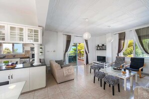 A modern living room with a sofa, dining table, and chairs. There is a kitchen area with cabinets and a window.