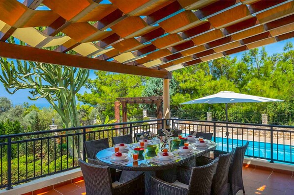 An outdoor dining area with a table set for six, surrounded by wicker chairs, overlooking a swimming pool and lush greenery.