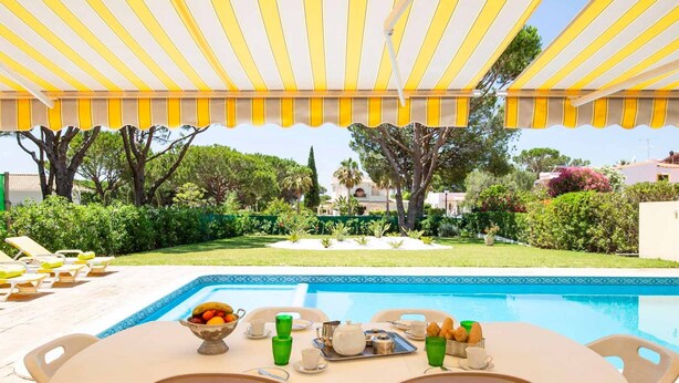 Un espace aménagé au bord de la piscine, avec une table dressée pour le petit-déjeuner, entouré de chaises longues et surmonté d’un parasol à rayures jaunes.