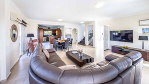 A modern living room with a large leather sofa, a flat-screen TV, and a dining area in the background.