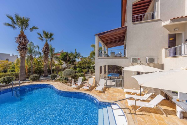 A swimming pool with lounge chairs and umbrellas, surrounded by palm trees and a building with balconies.