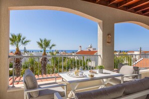 A balcony with a table set for dining, overlooking a coastal view with palm trees and a clear sky.