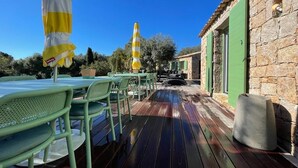 Un patio al aire libre con sillas y mesas de metal verde, un paraguas a rayas amarillo y blanco, y un edificio de piedra con contraventanas verdes.