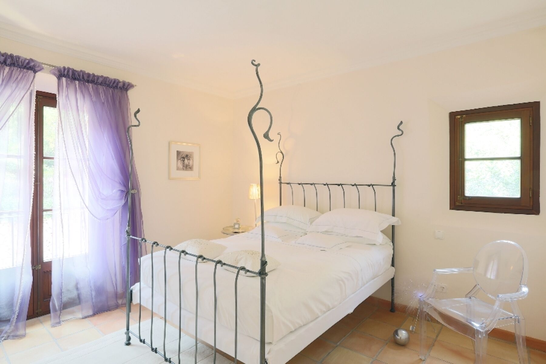 A bedroom with a metal bed frame, white bedding, a clear plastic chair, a small table with a lamp, and a picture on the wall.