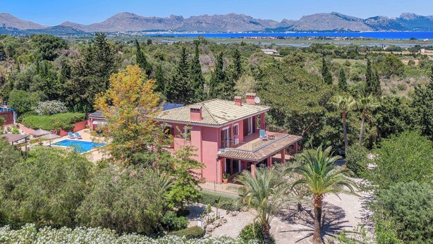 A pink villa with a pool, surrounded by trees and mountains in the background.