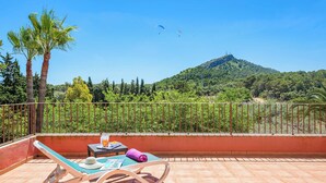 A balcony with a view of a hill, a table with a drink, and lounge chairs.