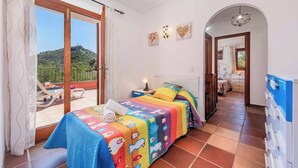 A bedroom with a colorful quilt, a nightstand with a lamp, a view of a balcony, and a hallway leading to another room.