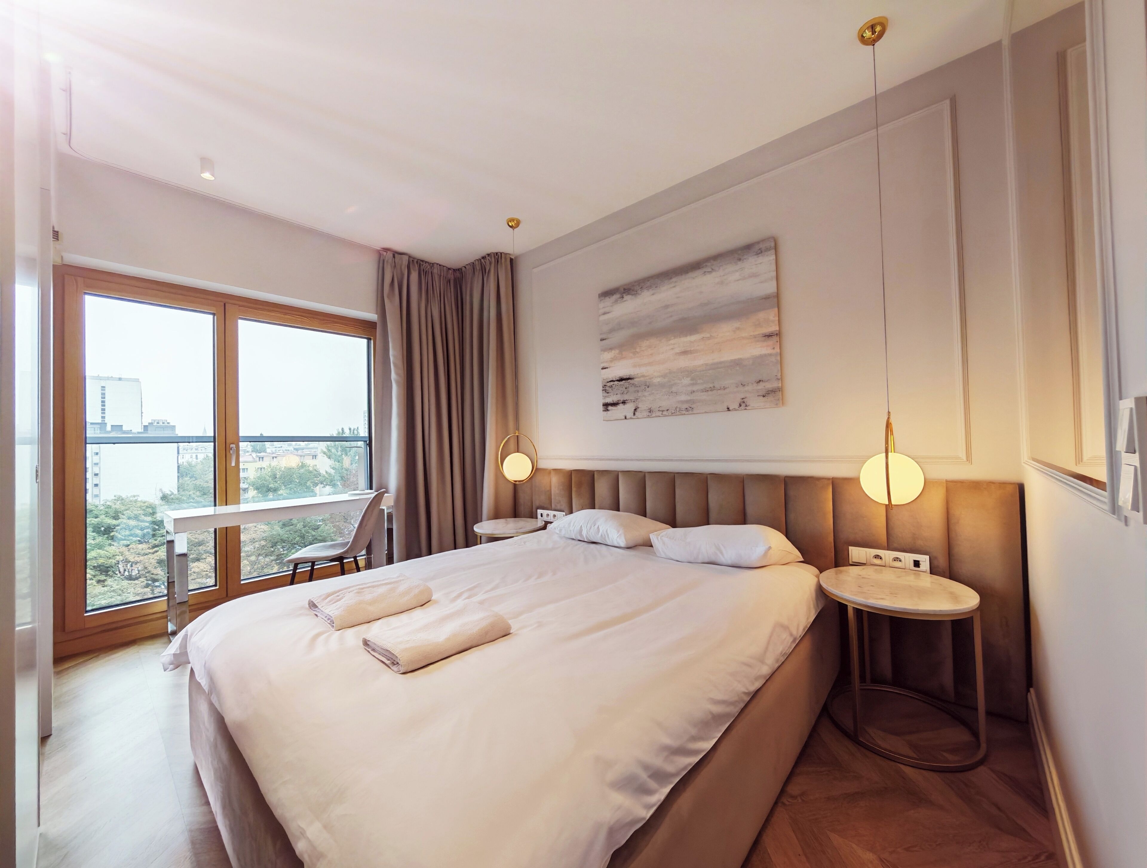 Ein Hotelzimmer mit einem großen Bett, Nachttischen, einem Schreibtisch und Blick auf die Stadt durch große Fenster.
