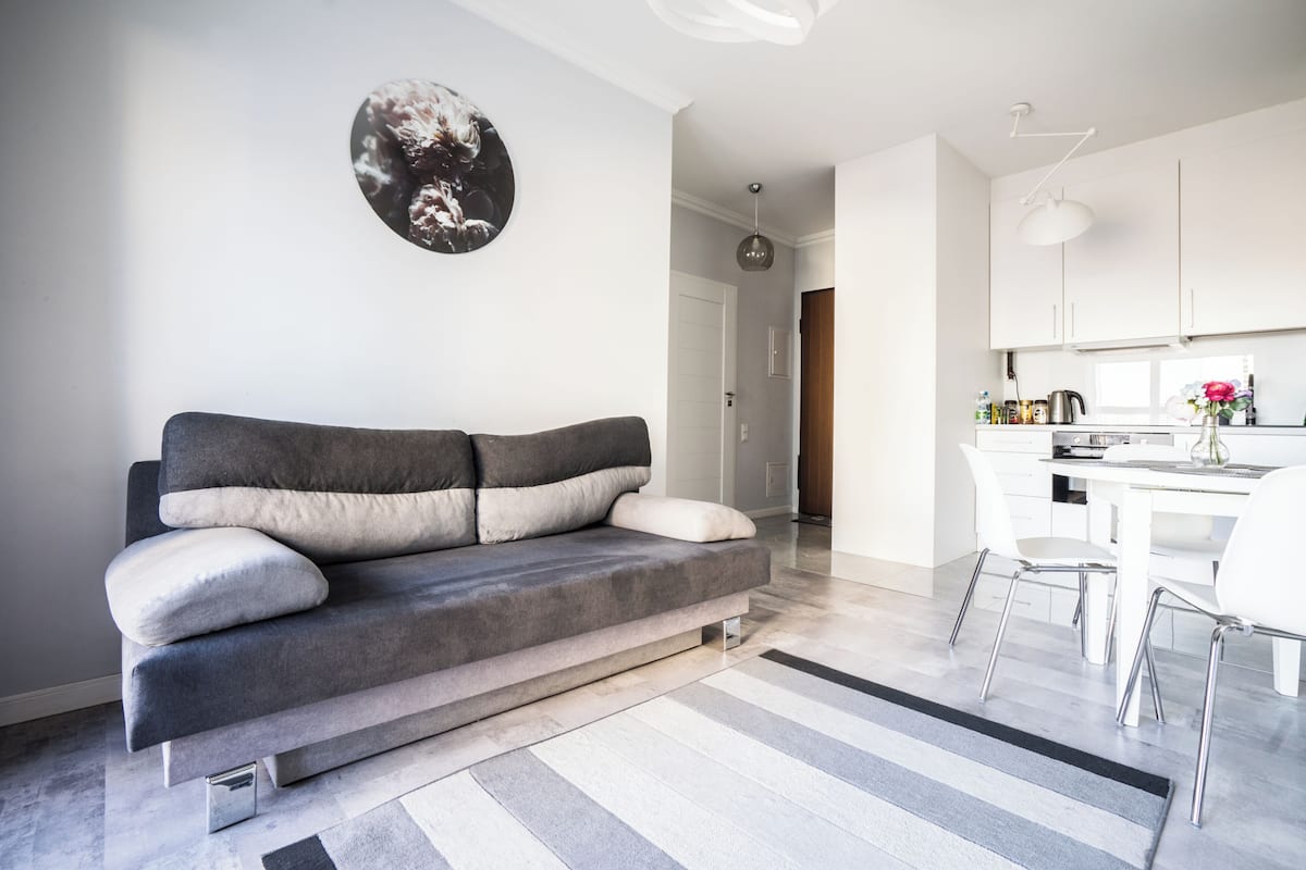 A modern living room with a grey sofa, a striped rug, and a kitchen area with white cabinets and a dining table.