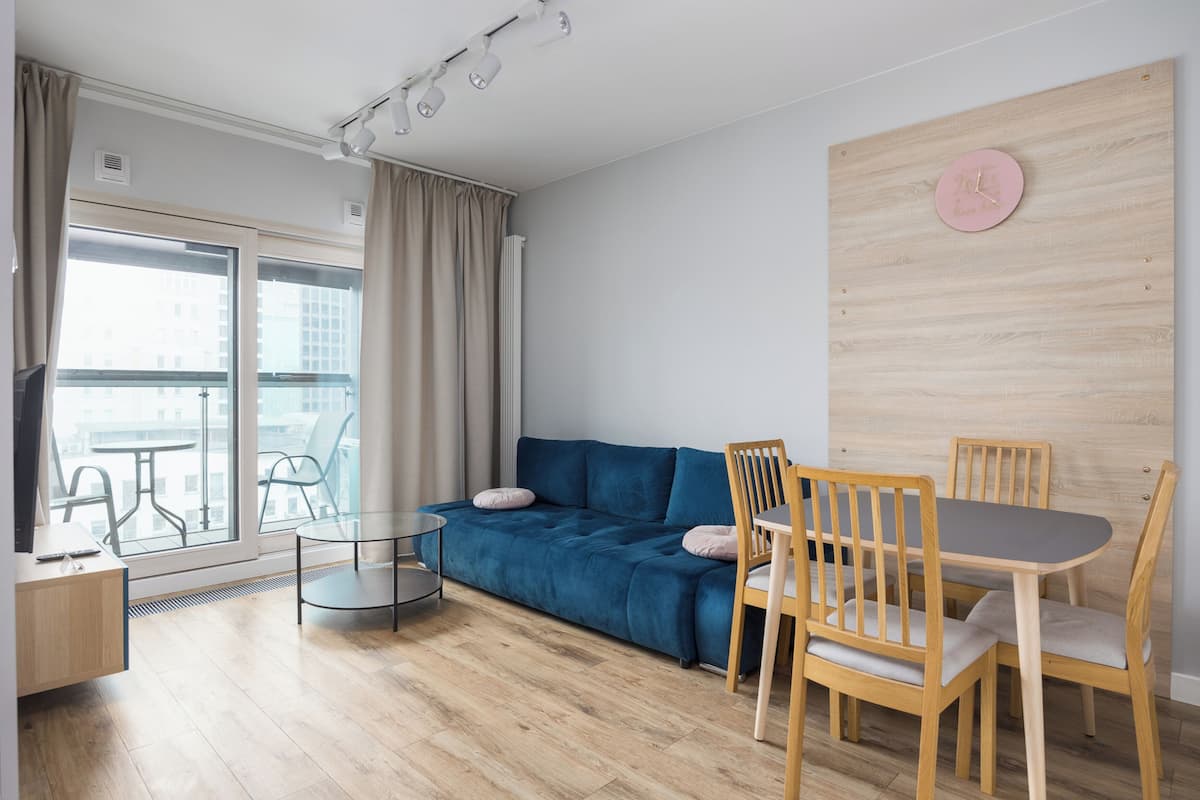 A modern living room with a blue sofa, wooden dining table, and chairs. There is a glass coffee table, a TV on a wooden cabinet, and a view through large windows.