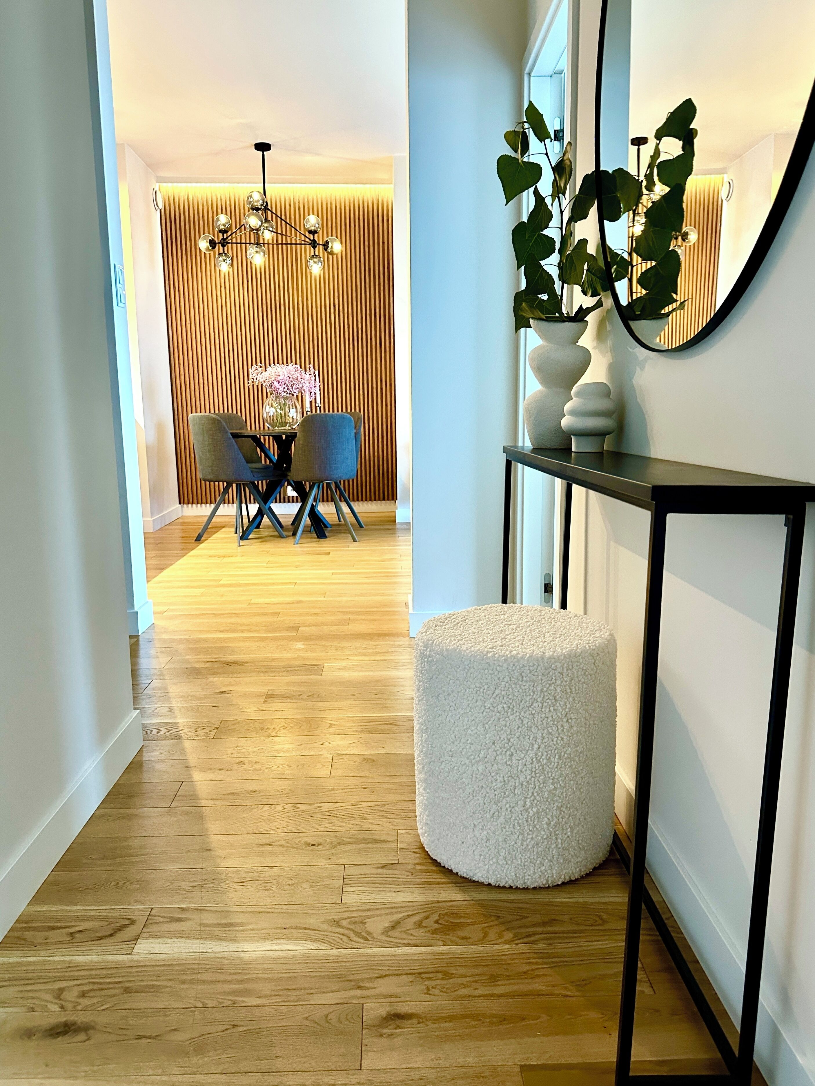 A modern interior with a wooden floor, a dining area with two chairs and a table, a white pouf, a console table with a mirror, and a potted plant.