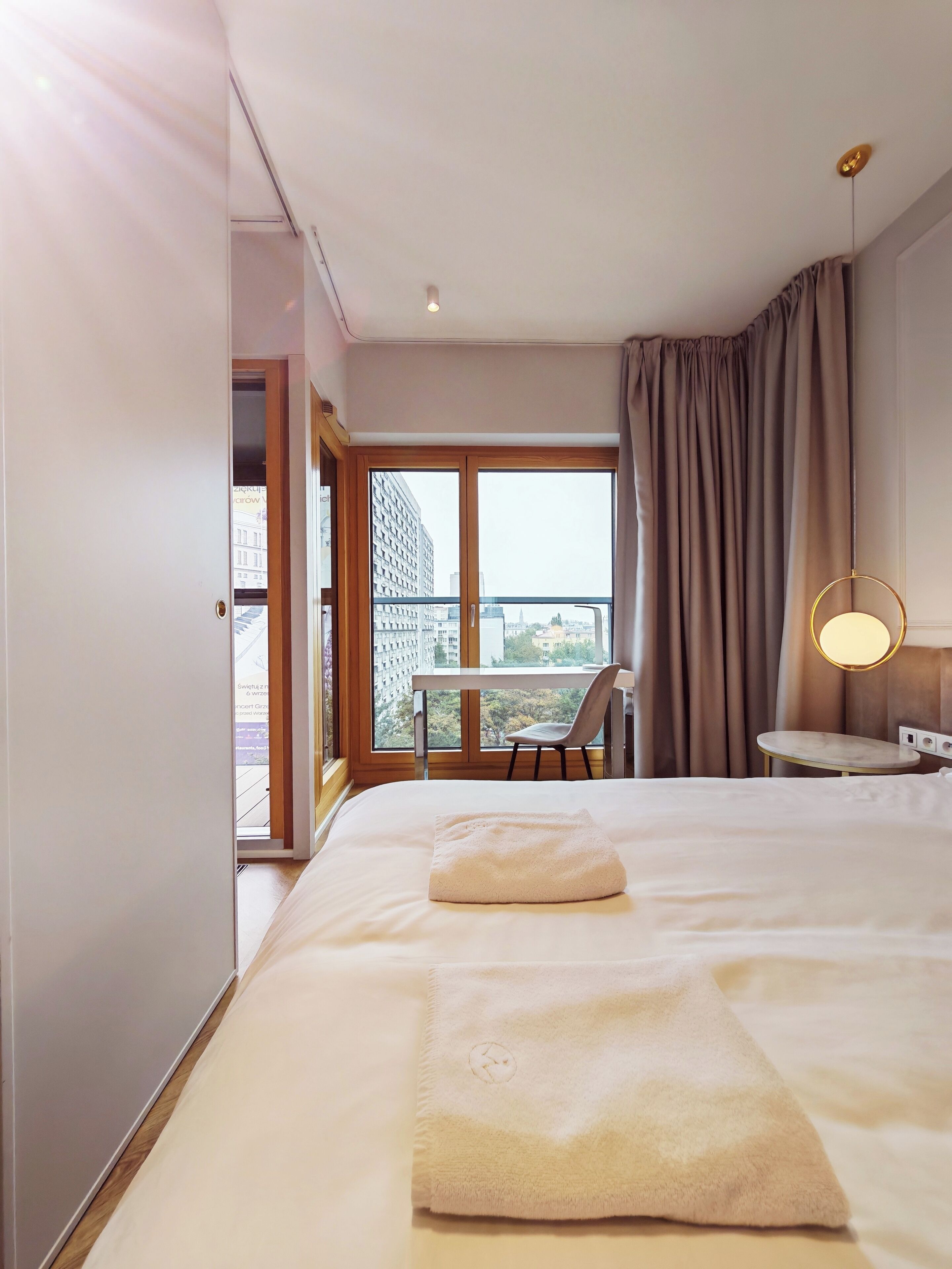 Ein Hotelzimmer mit einem Bett, einem kleinen Tisch, einem Stuhl, einem Balkon mit Blick auf Gebäude und einer Hängeleuchte.