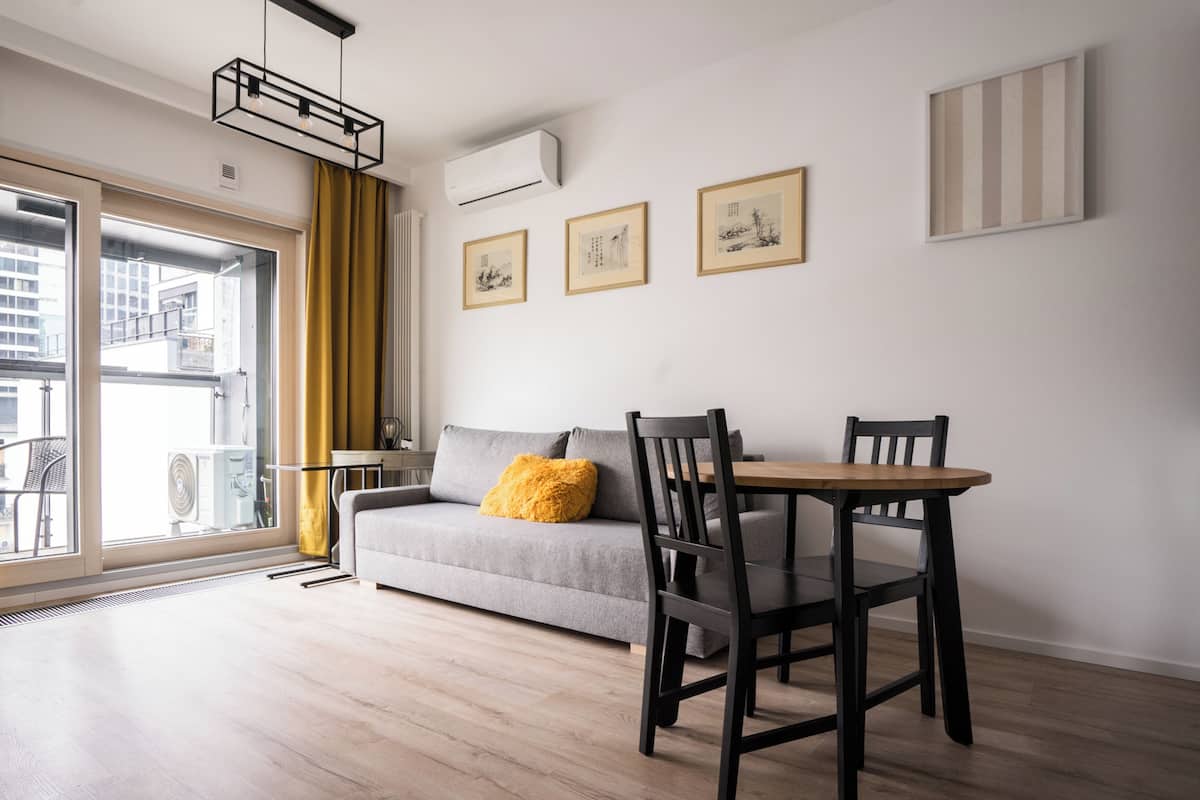 A modern living room with a grey sofa, a wooden dining table, and chairs. There are three framed pictures on the wall, a window with yellow curtains, and an air conditioning unit.