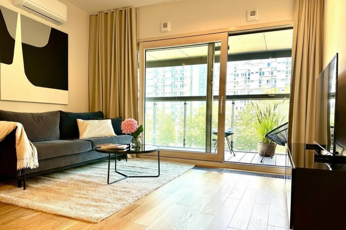 A modern living room with a sofa, coffee table, and a TV. There is a large window with a view of buildings and greenery outside.