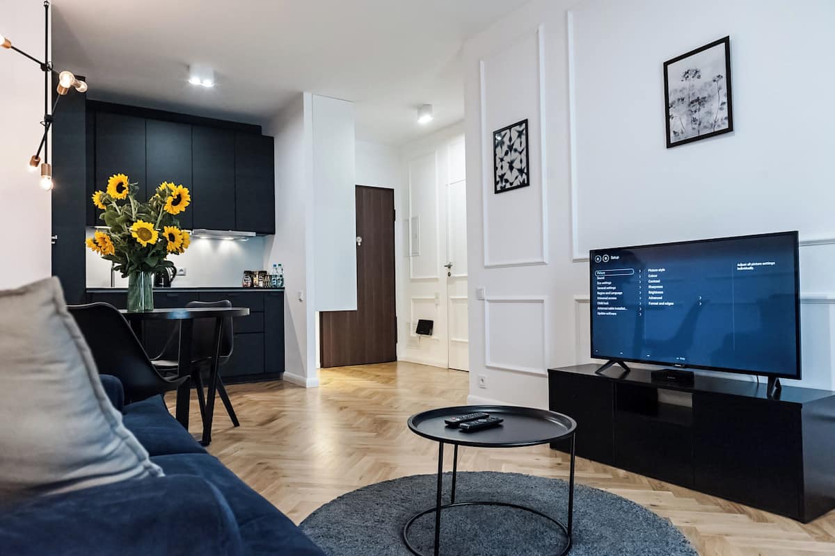 A modern living room with a sofa, a coffee table, and a TV. There is a kitchen area with dark cabinets and a dining table with a vase of sunflowers.