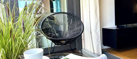 A room with a black chair, a white cup on a saucer, a magazine, and a plant with long leaves.