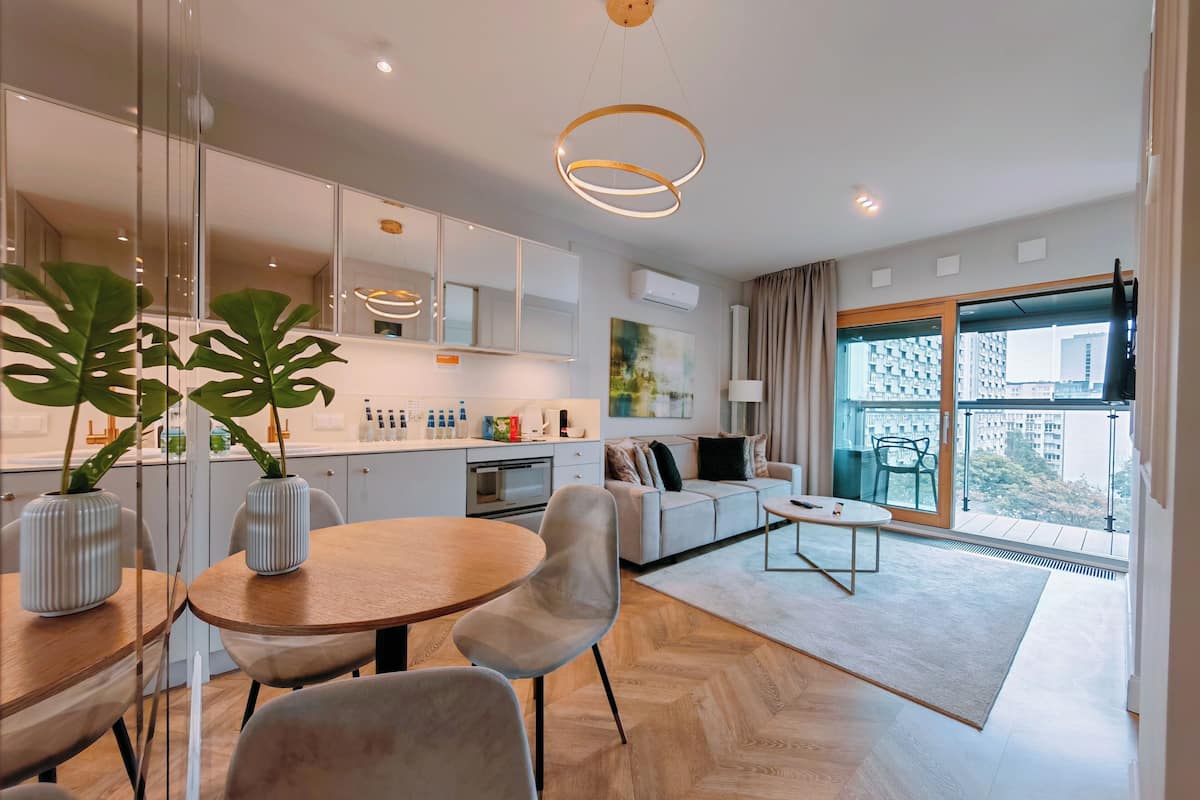 A modern apartment with a kitchen, dining area, and living room. It features a large window with a city view, a sofa, and a dining table with chairs.