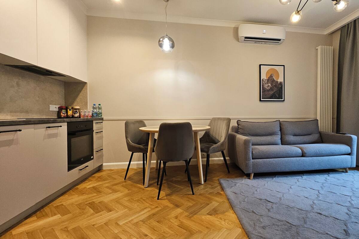 A modern living room with a grey sofa, a dining table with two chairs, a kitchen area with a stove and cabinets, and a large window with curtains.