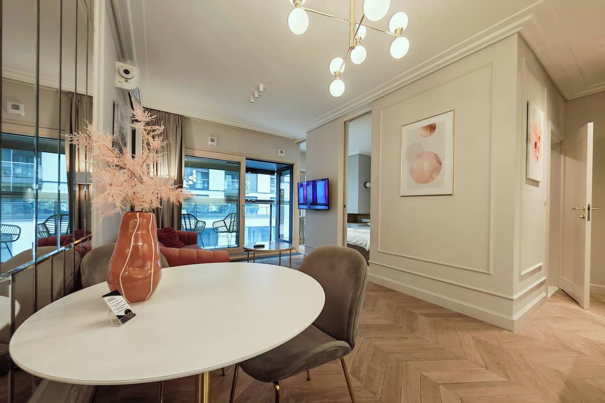 A modern hotel room with a round dining table, a vase with dried flowers, a sofa, and a large window offering a view of the city.