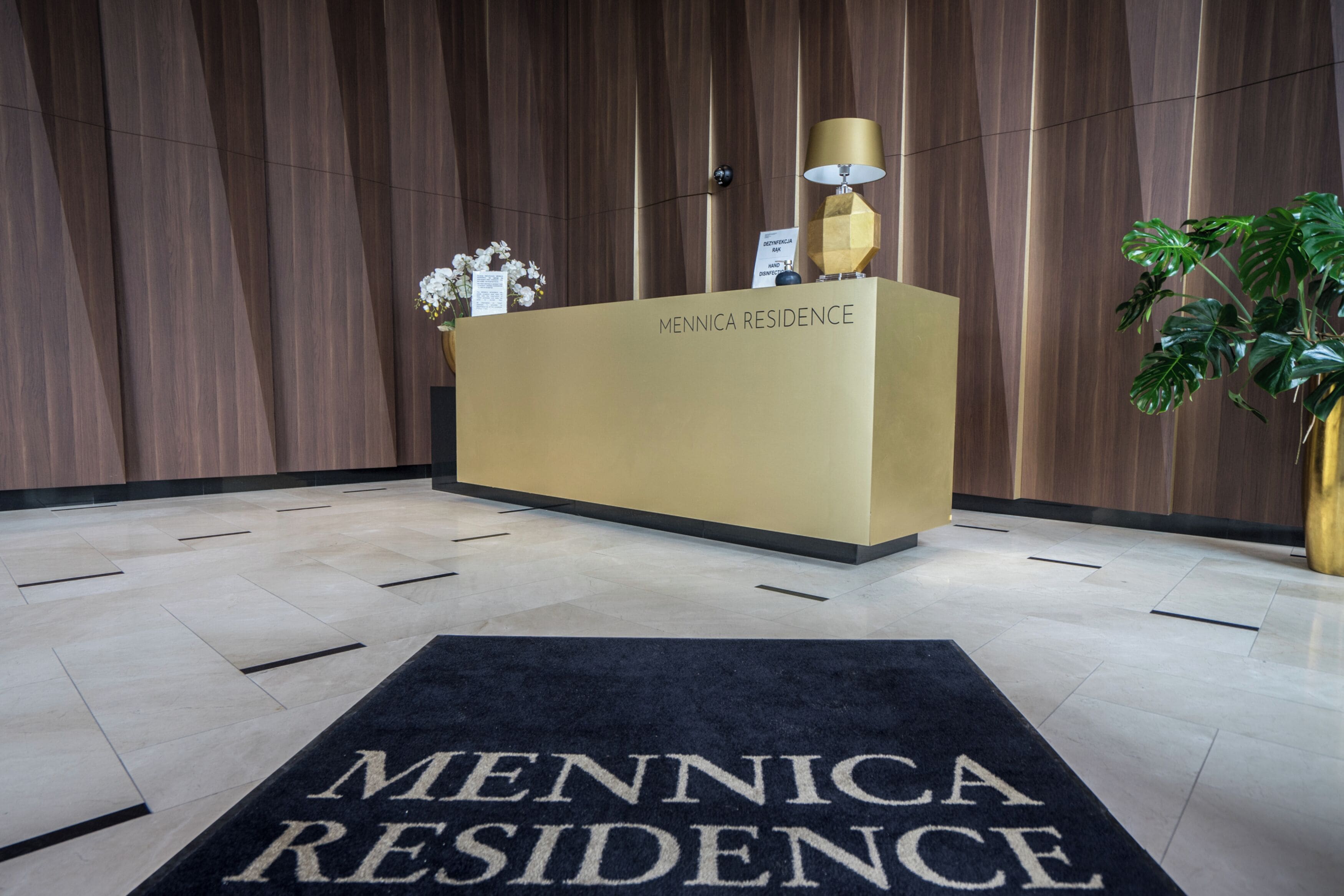 Ein Empfangstresen mit der Beschriftung 'Mennica Residence', einer Zimmerpflanze und einem schwarzen Teppich, ebenfalls mit diesem Namen.