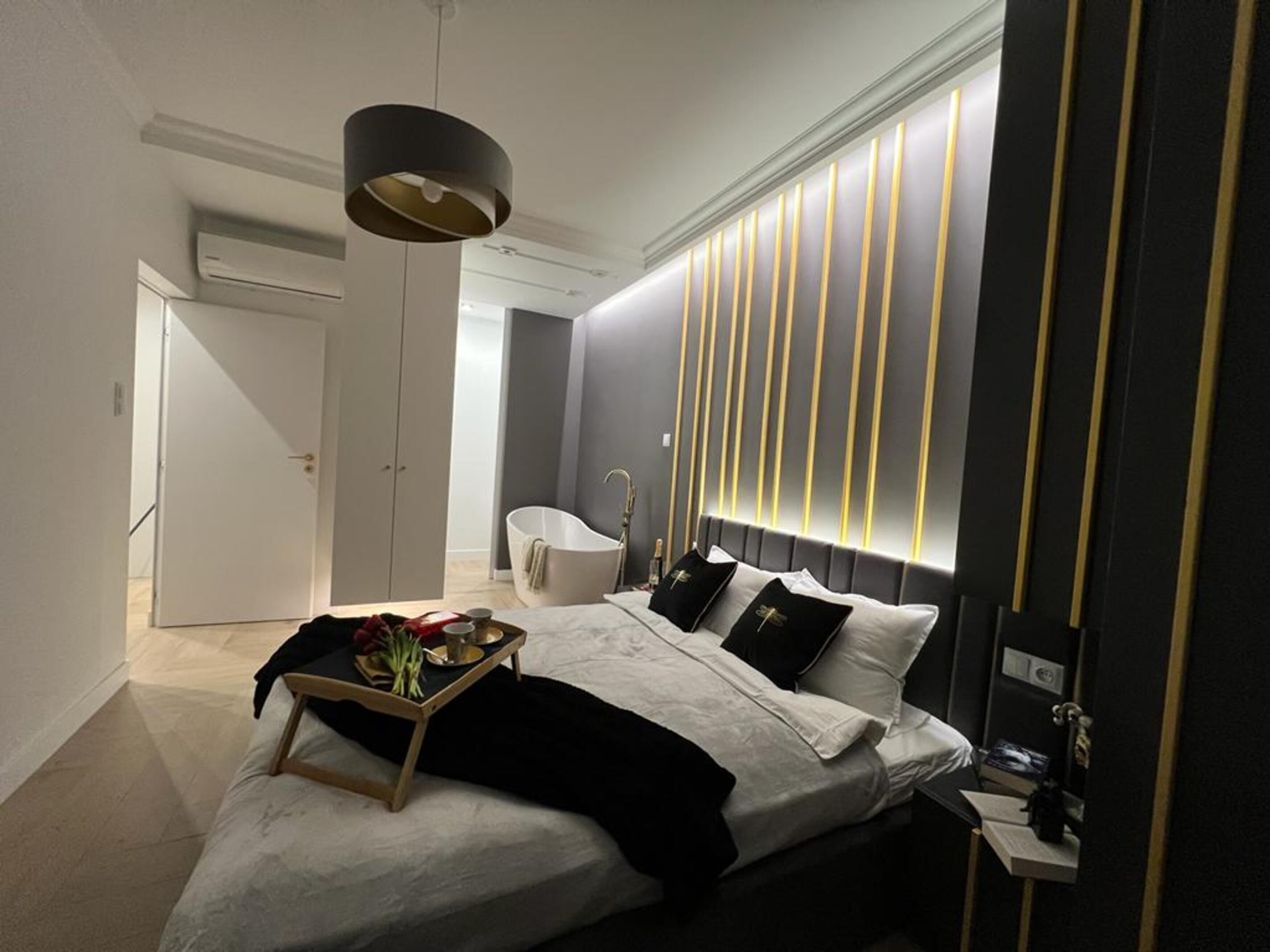 A modern bedroom with a large bed, bedside tables, a vanity, and a bathtub.