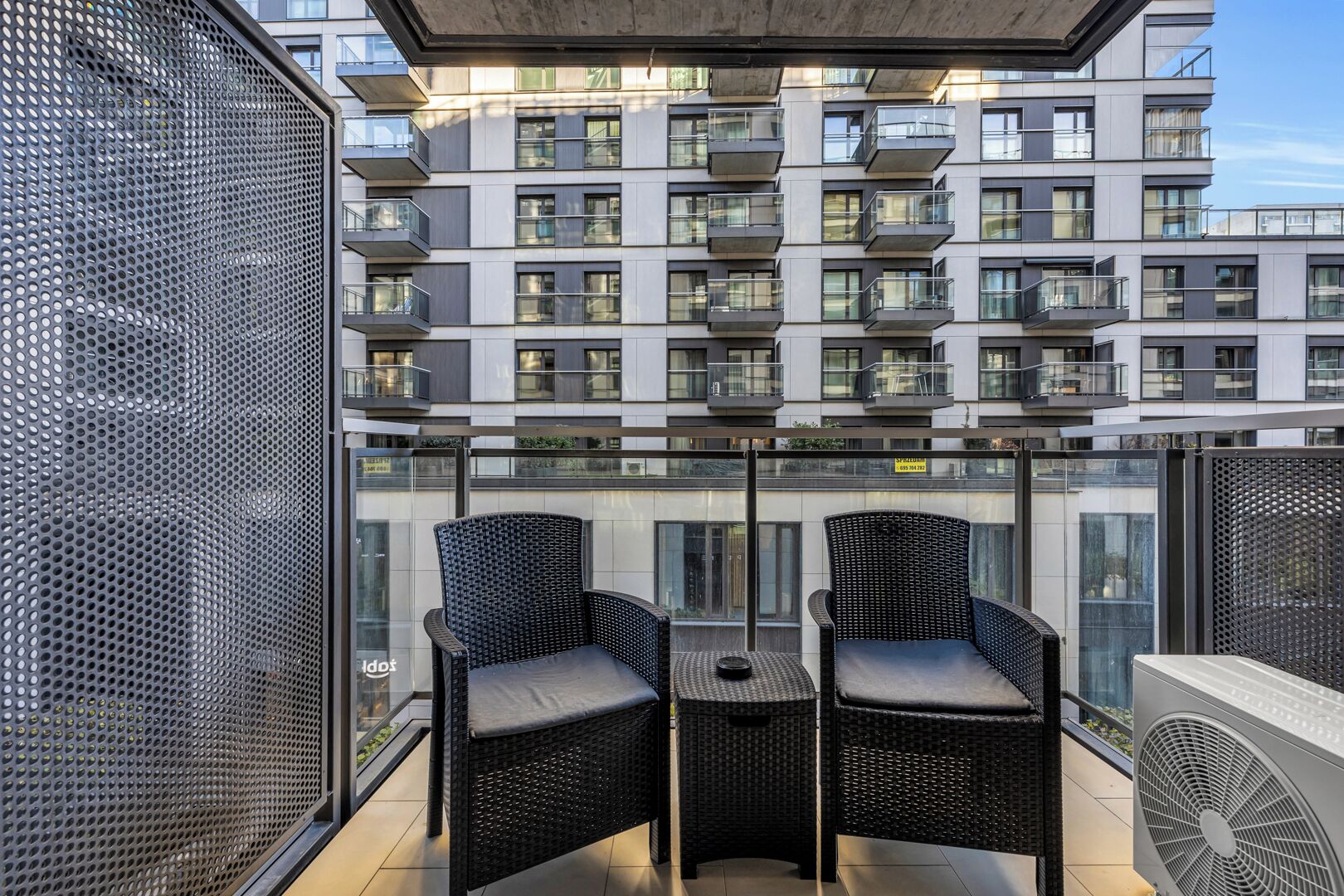 A balcony with two wicker chairs and a table, overlooking a modern apartment building with balconies.