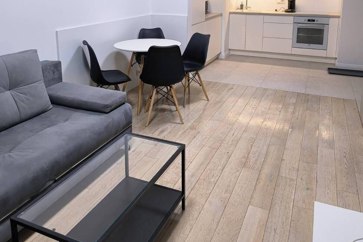 A modern apartment with a grey sofa, a dining table with chairs, a kitchen area with white cabinets, and a small dining table.