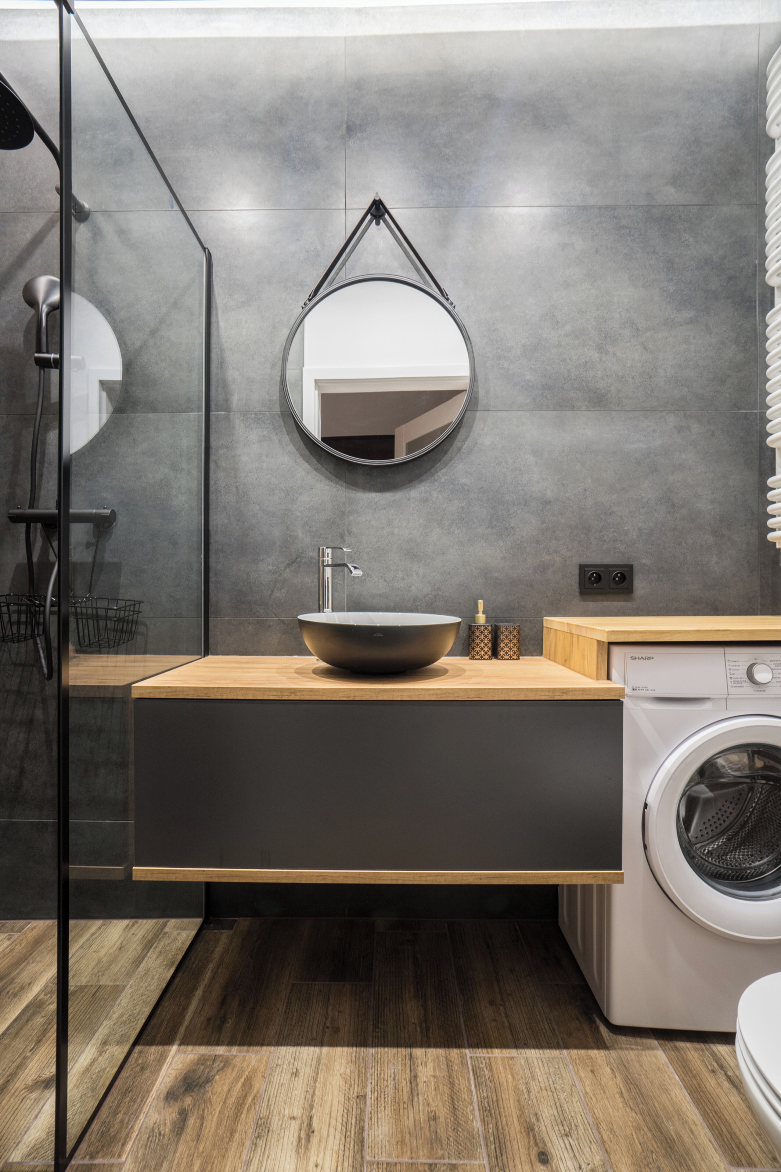Ein modernes Badezimmer mit einem Holzbadezimmer-Möbel, einem runden Spiegel, einem Waschbecken, einer Waschmaschine und einer Duschkabine aus Glas.