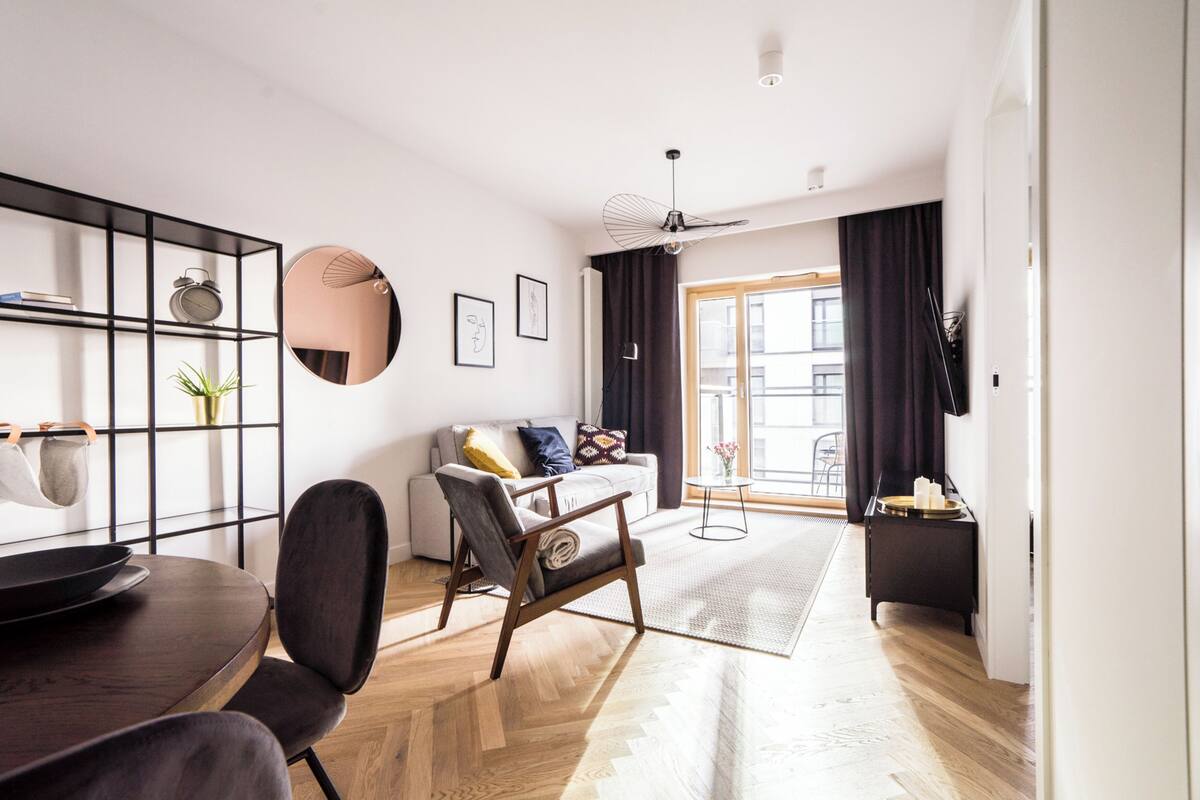 A modern living room with a sofa, a dining table, and a coffee table. There is a black bookshelf with a mirror and a plant. The room has wooden flooring and a large window with curtains.