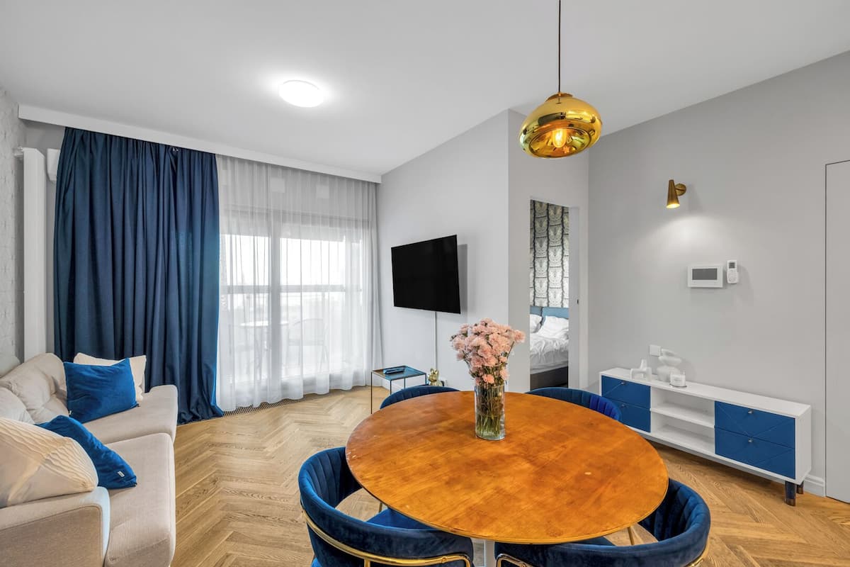 A modern living room with a round wooden table, blue upholstered chairs, a beige sofa with blue cushions, a flat-screen TV mounted on the wall, a bedside table with a lamp, and a window with sheer curtains.
