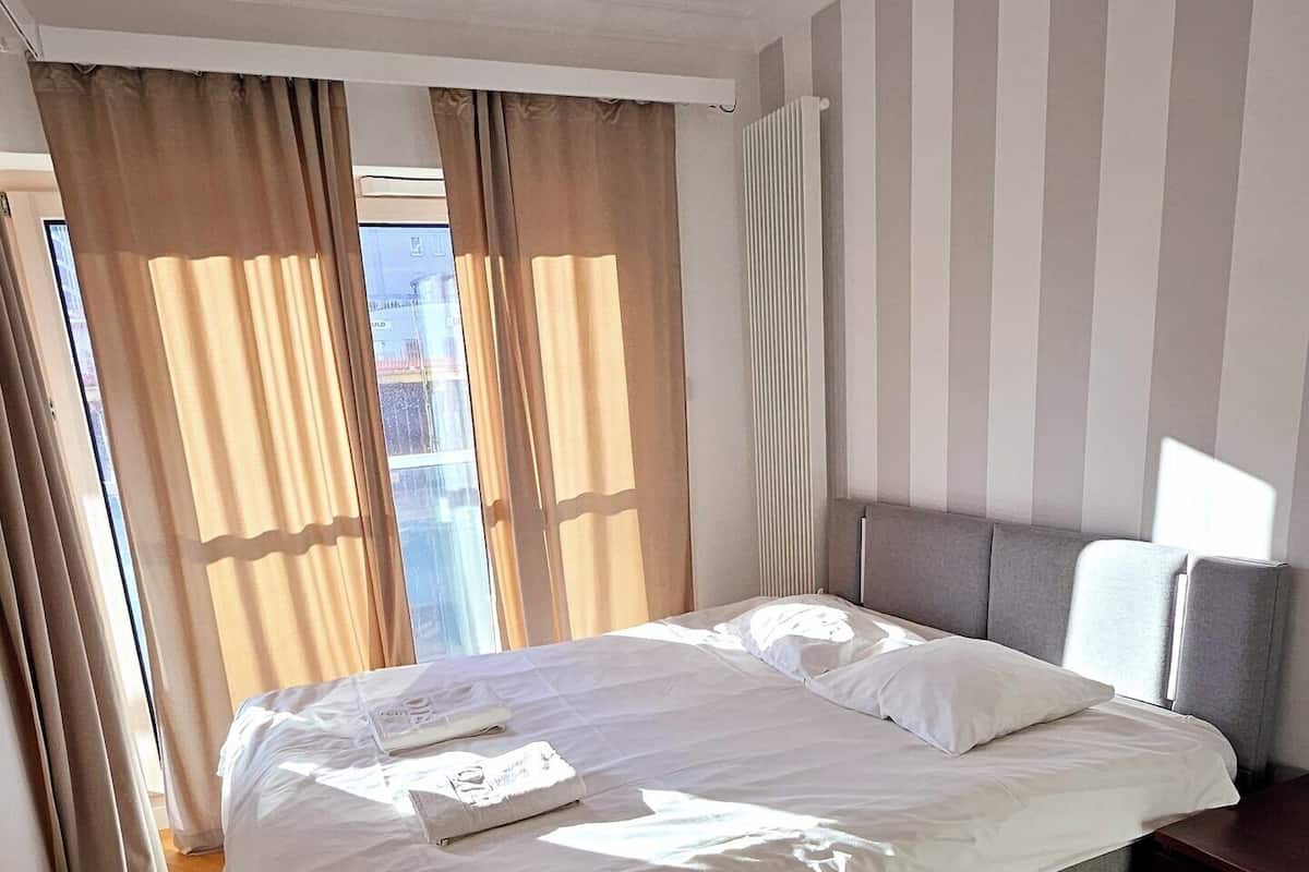 A modern hotel room with a large bed, wooden parquet flooring, and a view through the window.