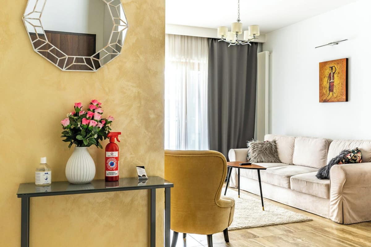A living room with a yellow accent wall, a round mirror, a vase with flowers, a spray bottle, a small table, a yellow armchair, a beige sofa, and a painting on the wall.