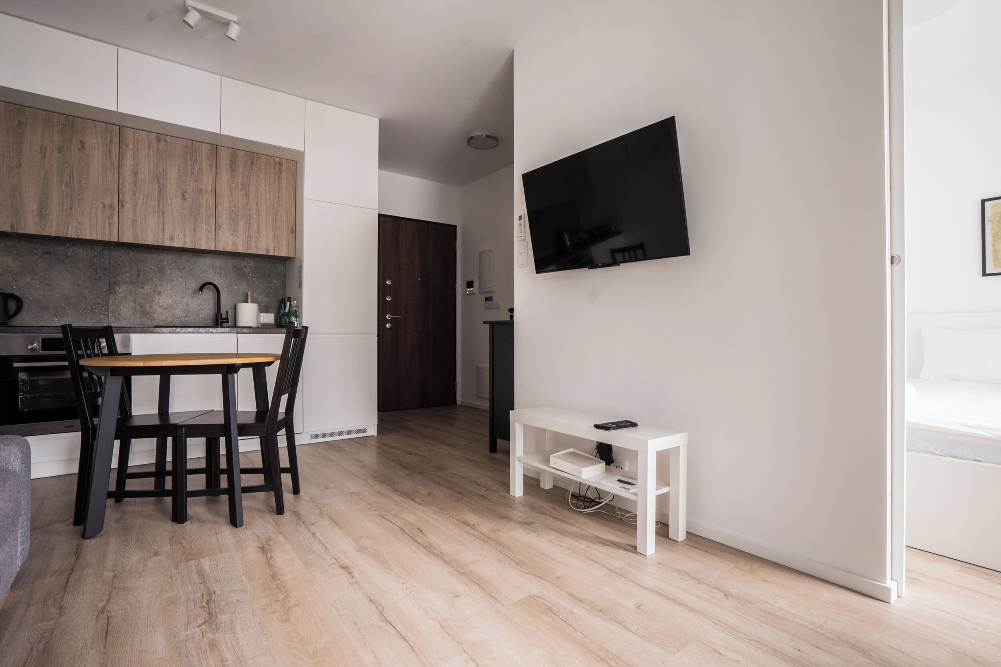A modern kitchen with wooden cabinets, a dining table with chairs, a white TV on the wall, and a small white side table.